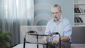 Old male sitting on couch and looking at walking frame standing in front of him