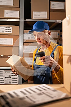 Old male warehouse worker scans cardboard box with Barcode Scanner. Rows of cardboard boxes with products for shipment