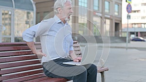 Old Man with Back Pain Using Laptop while Sitting Outdoor on Bench