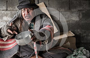 Old man beggar homeless is hair gray wears glasses and hat sitting on cardboard in shelter holding empty bowl for asks donate food