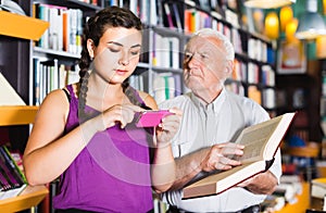 Old man is choosing book while girl chatting by phone in bookstore.