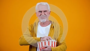 Old man displeased with uninteresting movie, holding popcorn, yellow background