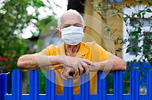 Old man in facem mask leaning on fence near his country house