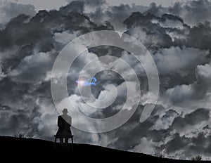 A old man in a fedora sits on a bench in front of a wall of storm clouds but a glimmer of sunlight shines through a small hole in