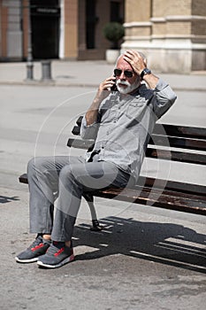 Old man forget something, speak on smartphone and hold hand on head, seating on bench,  stock photo