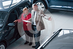 Old man in formal wear with notepad supporting woman in choosing automobile