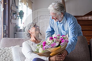 Old man giving flowers at his wife sitting on the sofa at home for the San Valentinesâ€™ day. Pensioners enjoying surprise