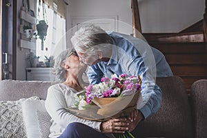 Old man giving flowers at his wife sitting on the sofa at home for the San Valentinesâ€™ day. Pensioners enjoying surprise kissing
