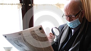 Old man in glasses and a black jacket is sitting in a chair by the window reading a fresh newspaper