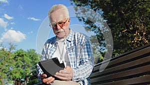 Old man with glasses and mustache is sitting on bench in park and reading ebook