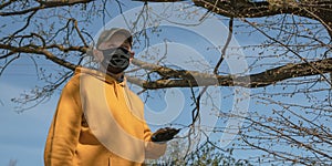 Old man in gloves and mask stands in garden at bare tree