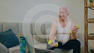 Old man in a good mood doing exercise at home while sitting on the sofa and watching something on the laptop he lifting