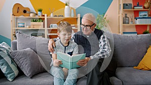 Old man grandfather reading book to smart grandchild indoors in apartment