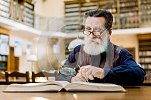 Old man with gray beard, in glasses, vintage clothes, reading a book in the ancient library, using magnifying glass