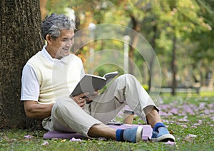 old man with grey hair sitting under a tree and reading a book in forest park