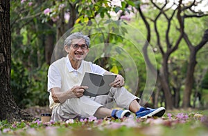 old man with grey hair wears eye glasses sitting under a tree and reading a book in forest park