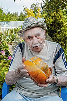 Old man in a hat with a pumpkin in the garden. gardener is holding a ripe orange pumpkin. concept of active old age