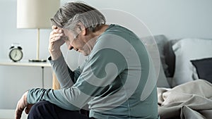 Old man holding his head with his hands sits on the bed