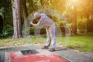 Old man at a minigolf court hits a ball with an iron racket. A w