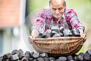 Old man picking up a basket full of coal briquettes from a pile in the backyard