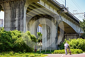 An old man pushes a bicycle and walks under a large railway bridge