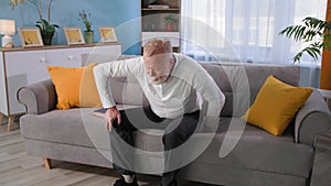 old man reads book then gets up from the couch and feels pain in his leg, granddad with osteoporosis rubs his knee at