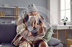 Old man is sitting at home, warmly wrapped in warm blanket, dressed in knitted sweater and hat.