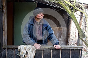 An old man in sloppy clothes stands on the threshold of his own ruined house and looks into the distance