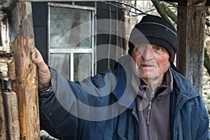 An old man in sloppy clothes stands on the threshold of his own ruined house and looks into the distance