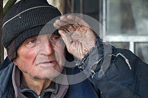 An old man in sloppy clothes stands on the threshold of his own ruined house and looks into the distance