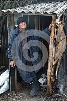 An old man in sloppy clothes stands on the threshold of his own ruined house and looks into the distance