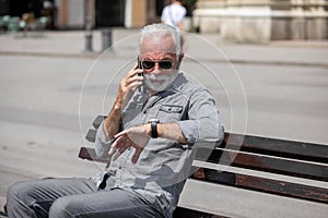 Old man speak on smartphone and looking on watch, seating on bench,  stock photo