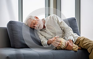 Old man suffering from knee pain lying on sofa in the living room, Elderly man suffering from headache while sitting on the sofa