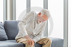 Old man suffering from knee pain sitting sofa in the living room, Elderly man suffering from knee pain while sitting on the sofa