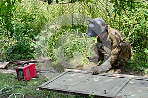 Old man welder in brown uniform, welding mask and welders leathers, weld metal door with arc welding machine outdoors