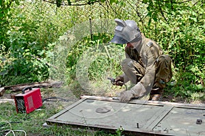 Old man welder in brown uniform, welding mask and welders leathers, weld metal door with arc welding machine outdoors