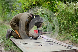 Old man welder in brown uniform, welding mask and welders leathers, weld metal door with arc welding machine outdoors