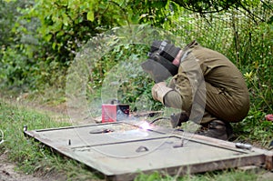 Old man welder in brown uniform, welding mask and welders leathers, weld metal door with arc welding machine outdoors