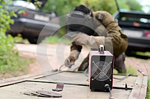 Old man welder in brown uniform, welding mask and welders leathers, weld metal door with arc welding machine outdoors