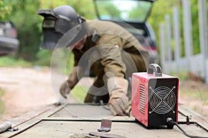 Old man welder in brown uniform, welding mask and welders leathers, weld metal door with arc welding machine outdoors