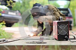 Old man welder in brown uniform, welding mask and welders leathers, weld metal door with arc welding machine outdoors