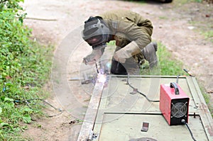 Old man welder in brown uniform, welding mask and welders leathers, weld metal door with arc welding machine outdoors