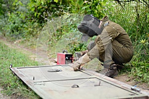 Old man welder in brown uniform, welding mask and welders leathers, weld metal door with arc welding machine outdoors