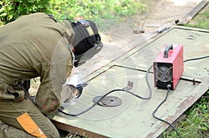 Old man welder in brown uniform, welding mask and welders leathers, weld metal door with arc welding machine outdoors