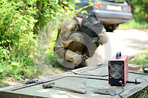 Old man welder in brown uniform, welding mask and welders leathers, weld metal door with arc welding machine outdoors