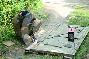 Old man welder in brown uniform, welding mask and welders leathers, weld metal door with arc welding machine outdoors