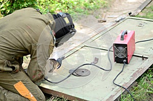 Old man welder in brown uniform, welding mask and welders leathers, weld metal door with arc welding machine outdoors