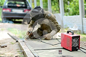 Old man welder in brown uniform, welding mask and welders leathers, weld metal door with arc welding machine outdoors