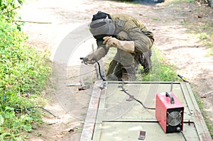 Old man welder in brown uniform, welding mask and welders leathers, weld metal door with arc welding machine outdoors