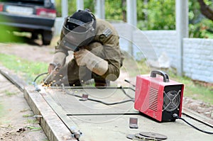 Old man welder in brown uniform, welding mask and welders leathers, weld metal door with arc welding machine outdoors